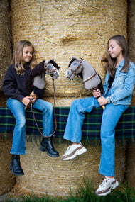 Zwei Mädchen sitzen auf Strohballen auf einer karierten Decke und halten jeweils ein Hobby Horse Guccini von Hobby Horse LarDen – grauer Kopf im Holsteiner-Stil mit weißem Nasenstreifen und langer dunkelbrauner Garnmähne – mit dem fellgefütterten Halfter Walnut, dem Brustgurt Walnut mit Fell und dem Führstrick Vivaldi