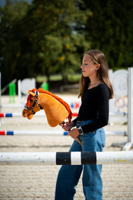 Mädchen im schwarzen Oberteil steht hinter einer Sprungstange auf einem Sandplatz und hält das Hobby Horse Little Angel mit schwarzem Halfter von Hobby Horse LarDen – Fuchs-Plüschkopf, leuchtend rote Garnmähne – hier mit dem Espresso Fellhalfter aus Kunstleder