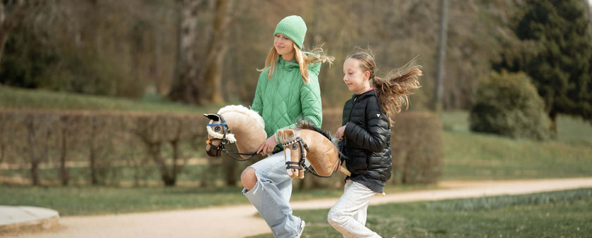 Two girls are enjoying hobby horsing outside, riding their toy hobby horses. One wears a green hat and jacket, the other a black jacket. They appear to be running or trotting along a path in a park or garden.