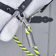 Gros plan sur un fermoir métallique reliant une sangle noire aux Hobby Horse - Rênes rayures citron jaunes, noires et grises de Hobby Horse LarDen. L’arrière-plan est en bois gris flou avec une partie d’un objet blanc doux visible.