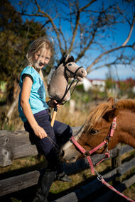 Lifestyle photo: a blonde girl in a turquoise top sits on a wooden paddock fence holding the Hobby Horse Piccolino with Black Halter by Hobby Horse LarDen - light beige plush head with a dark yarn mane - a chestnut pony behind her, shown here with the Complete black bridle from leatherette.