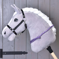 Hobby Horse LarDen „Halsring Streifen lila“ ist ein graues Hobby Horse mit weißer Mähne, schwarzem Zaumzeug und handgefertigtem lila Polypropylen-Halsring an einem Holzstab, abgebildet vor einer Wand aus Holzplanken.