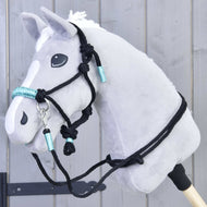 Das Hobby Horse LarDen Seilset Turquoise ist an einem grauen plüschigen Hobby Horse mit weißer Mähne, schwarzem Zaumzeug und türkisen Akzenten auf einem Holzstab vor einer grauen Holzwand zu sehen.