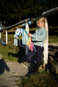 Mädchen im mintfarbenen Sweatshirt steht an einer weißen Begrenzung neben dem Reitplatz und hält das Hobby Horse Rainbow New von Hobby Horse LarDen mit weißem Plüschkopf und langer pastellfarbener regenbogenfarbener Garnmähne, hier mit dem Halfter blau mit Fell und dem Führstrick stripe blau