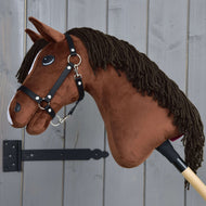 Das Hobby Horse LarDen „Hobby Horse Rocky mit Schwarzem Halfter“, ein braunes Plüsch-Steckenpferd, ist neben einer grauen Holzwand zu sehen.