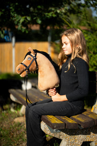 Mädchen in schwarzem Pullover sitzt in der Abendsonne auf einer Parkbank und hält das Hobby Horse Samantha mit schwarzem Halfter von Hobby Horse LarDen – sandfarbener Plüschkopf, schwarze Garnmähne – mit schwarzem Zaumzeug und Riemenzügeln