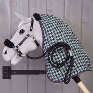 Set Harmony Green von Hobby Horse LarDen – ein Plüsch-Hobby Horse mit schwarzem Halfter für Hobby Horse, grün-violetter Stalldecke mit Hahnentrittmuster und aufgerolltem schwarzem Seil, an einer hölzernen Wandhalterung vor einer grauen Holzwand.