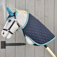 Hobby Horse LarDen Set Supreme Turquoise mit grauem Plüsch-Steckenpferd mit blauen Ohren, schwarzem Zaumzeug und gesteppter dunkelblauer Decke mit türkisfarbener Einfassung, auf einer Holzstange vor einer hellen Holzwand.