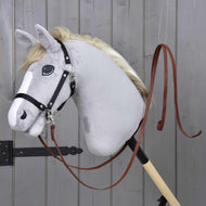 Ein graues plüschiges Hobby Horse von Hobby Horse LarDen mit schwarzem Zaumzeug und Offene Westernzügel Dark Brown ist auf einem Holzstab montiert, vor einer grauen Holzwand.