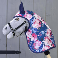 Das Hobby Horse LarDen „Regendecke Pfingstrose“ ist ein graues Hobby Horse mit schwarzem Zaumzeug, das eine blaue Regendecke mit rosa und weißen Blumen trägt, montiert auf einem Holzstab vor einer grauen Holzwand.