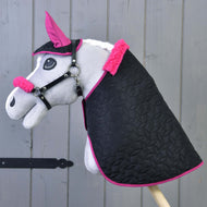Das Hobby Horse LarDen Set Thermo Pinkie (Ohrenkappe + Decke + Halfter mit Fell) enthält ein graues Hobby Horse mit einer schwarzen Decke mit pinker Einfassung, passender pinker Ohrenkappe und Halfter für Hobby Horse mit Fell, vor Holz-Hintergrund.