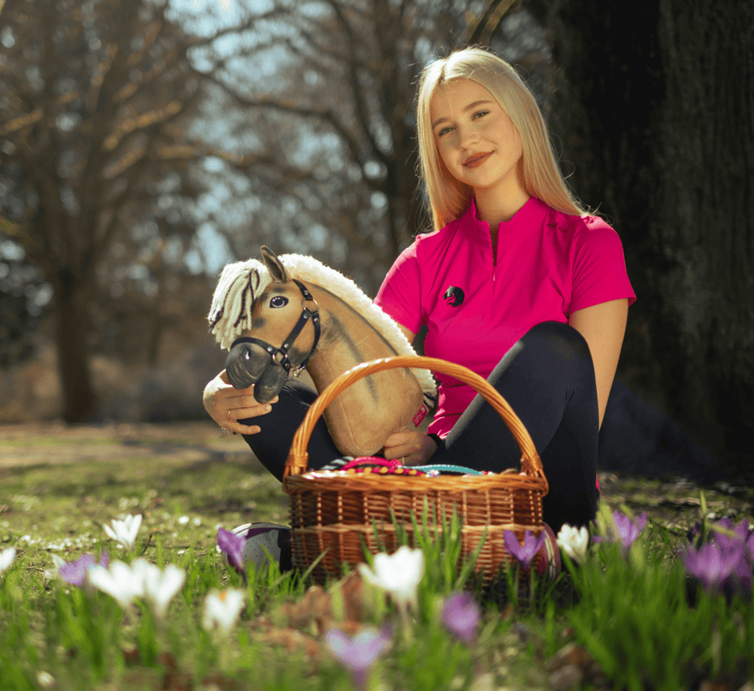 Jeune femme en chemise rose vif assise sur l’herbe en extérieur, tenant un jouet Hobby Horse et un panier en osier rempli d’œufs colorés, entourée de fleurs blanches et violettes en fleurs.