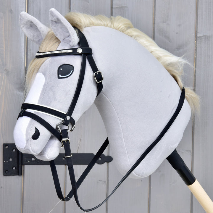 Ein Plüsch-Hobby Horse mit weißer Mähne, schwarzem Zaumzeug und weißer Blesse im Gesicht ist an einem Holzstab befestigt und vor einem grauen Holz-Hintergrund positioniert.