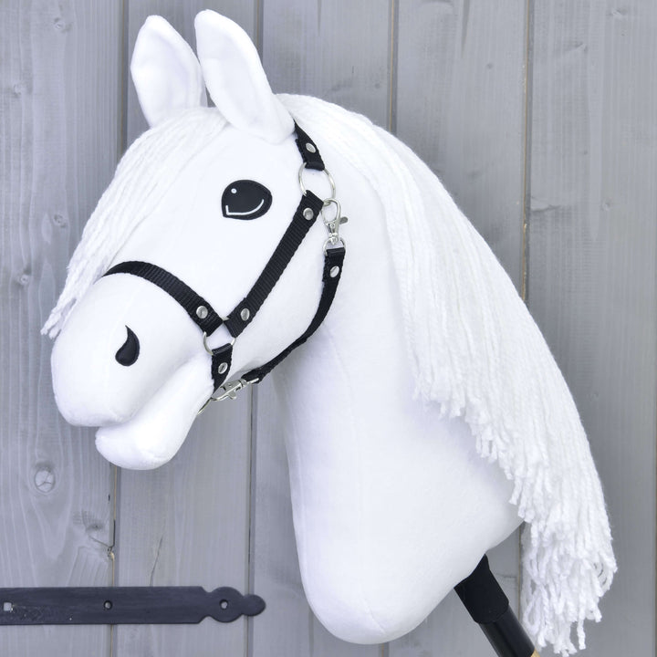 Ein weißes Hobby Horse mit schwarzem Zaumzeug und langer weißer Garnmähne auf einem Stock vor einer grauen Holzwand.