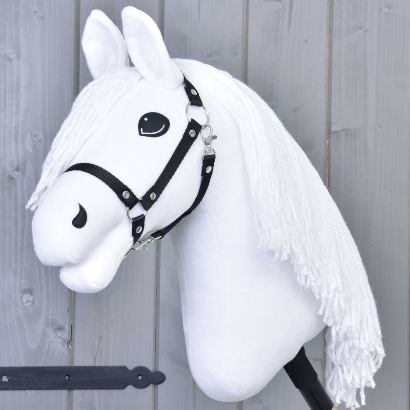Ein weißes Hobby Horse mit schwarzem Zaumzeug und langer weißer Garnmähne auf einem Stock vor einer grauen Holzwand.