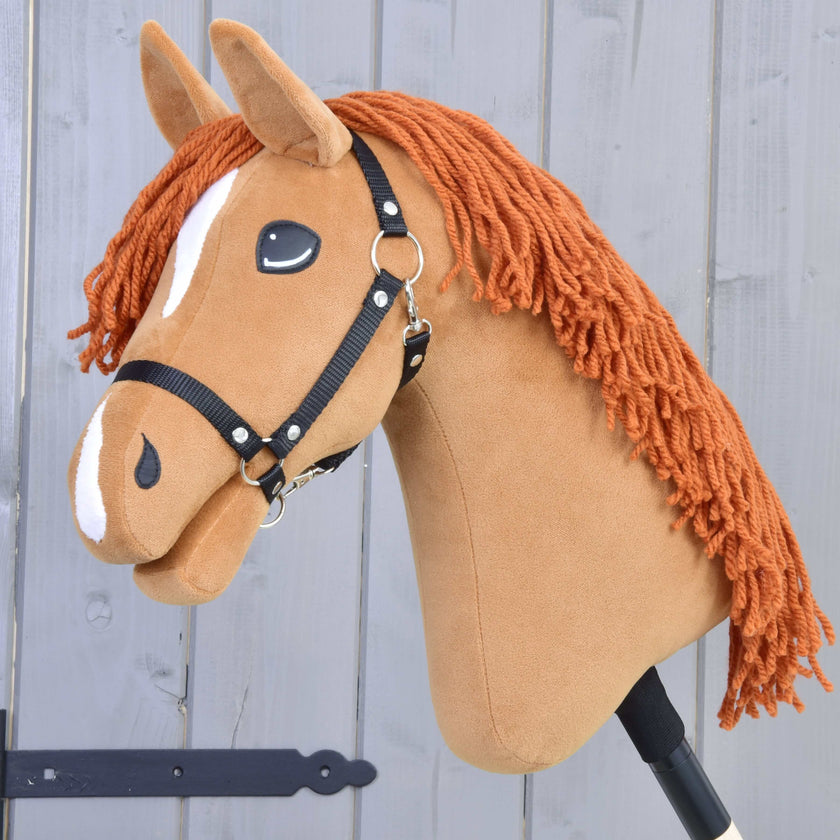 Ein handgefertigtes Hobby Horse mit braunem Stoffkopf, weißen Abzeichen, schwarzem Zaumzeug und orangefarbener Garnmähne auf einem Stock vor grauem Holz-Hintergrund.