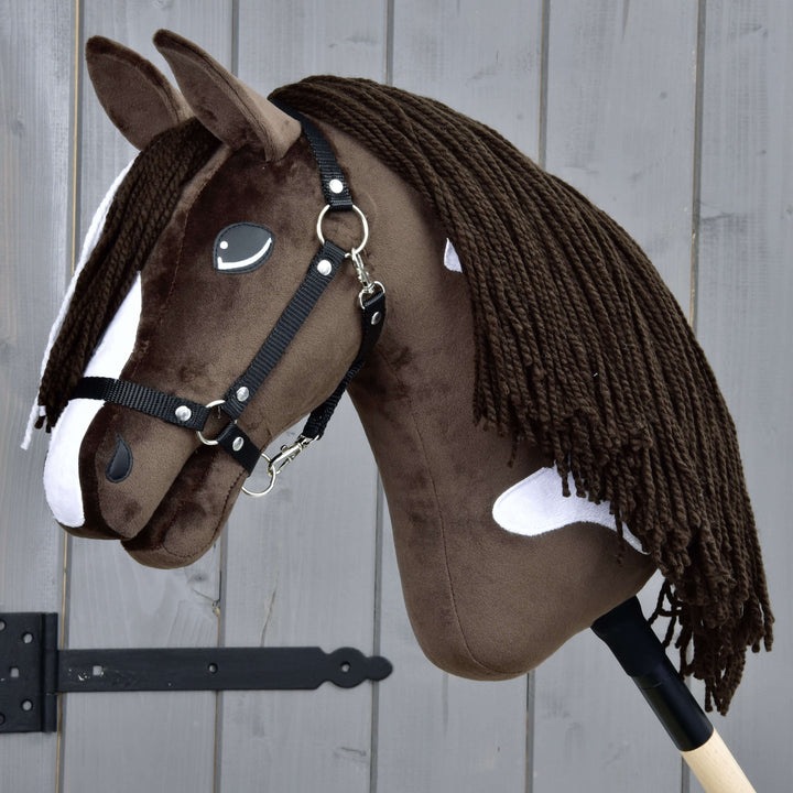 Ein realistisches braun-weißes Hobby Horse mit schwarzem Zaumzeug und langer Garnmähne auf einem Holzstock vor einem grauen Holzzaun.