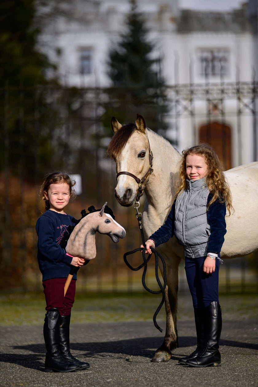 Zwei junge Mädchen stehen draußen neben einem weißen Pferd; ein Mädchen hält ein Spielzeug-Hobby Horse und zeigt damit ihre Liebe zu Hobby Horsing, während das andere die Zügel des echten Pferdes hält. Im Hintergrund sind ein unscharfes Gebäude und Bäume zu sehen.