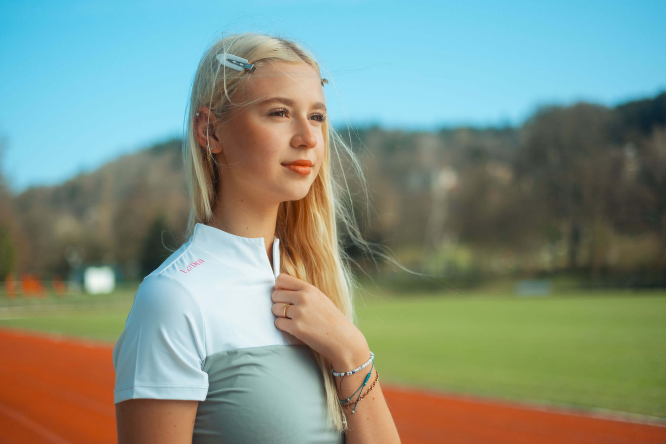 Eine junge blonde Frau in sportlichem Outfit steht auf einer Laufbahn im Freien und hält nachdenklich ihr Hobby Horse. Im Hintergrund sind grünes Gras, Bäume und ein blauer Himmel zu sehen.
