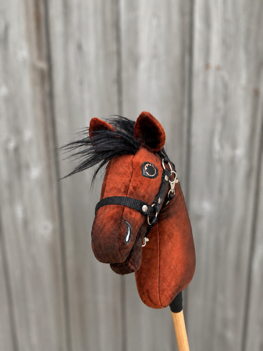 Hobby Horse LarDen's Hobby Horse Benny with Black Halter - size XS, a brown plush hobby horse with black mane and wooden stick handle, is pictured in front of a weathered wooden fence background.