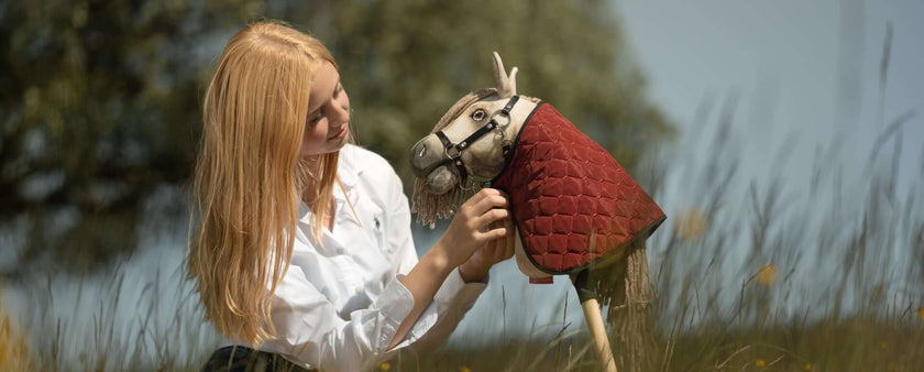 A young person with long blonde hair, wearing a white shirt, sits in tall grass outdoors and smiles while holding a Hobby Horse with a red quilted cover.