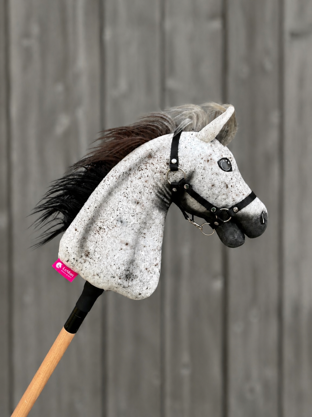 The Hobby Horse LarDen "Hobby Horse Luna with Black Halter- size S" features a gray and black Appaloosa-style horse head on a wooden stick, complete with bridle, reins, and a pink neck tag, pictured against gray wooden planks.