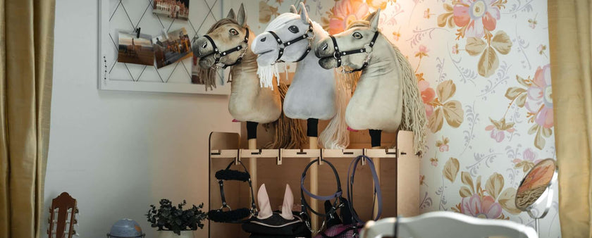 Three plush Hobby Horse heads are displayed on a wooden rack in a cozy, well-lit room with floral wallpaper. Below them hang riding accessories, celebrating the creative world of Hobby Horsing, while a corkboard with photos decorates the wall.