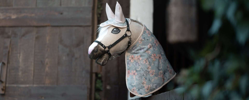 A realistic horse mannequin, perfect for hobby horsing enthusiasts, wears a floral-patterned hood and bridle, positioned to look out from a stable doorway.