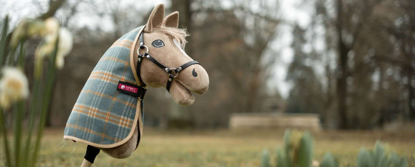 A fabric Hobby Horse with a plaid-patterned cloth and bridle stands upright outdoors on a stick, surrounded by grass and blurred trees in the background. Some white flowers are visible in the foreground—perfect for hobby horsing adventures.