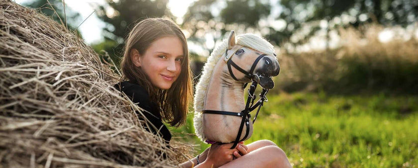 A young girl with brown hair sits by a haystack outdoors, smiling and holding her Hobby Horse with a white mane, as she enjoys Hobby Horsing in a sunlit grassy field.