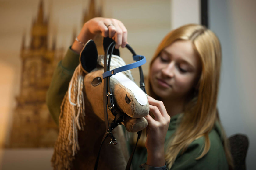 Ein junges Mädchen mit blonden Haaren legt ihrem Hobby Horse, einem Plüschspielzeug mit beiger Mähne, ein Zaumzeug an. Sie ist konzentriert und lächelt leicht in einer sanft verschwommenen Innenraumkulisse.