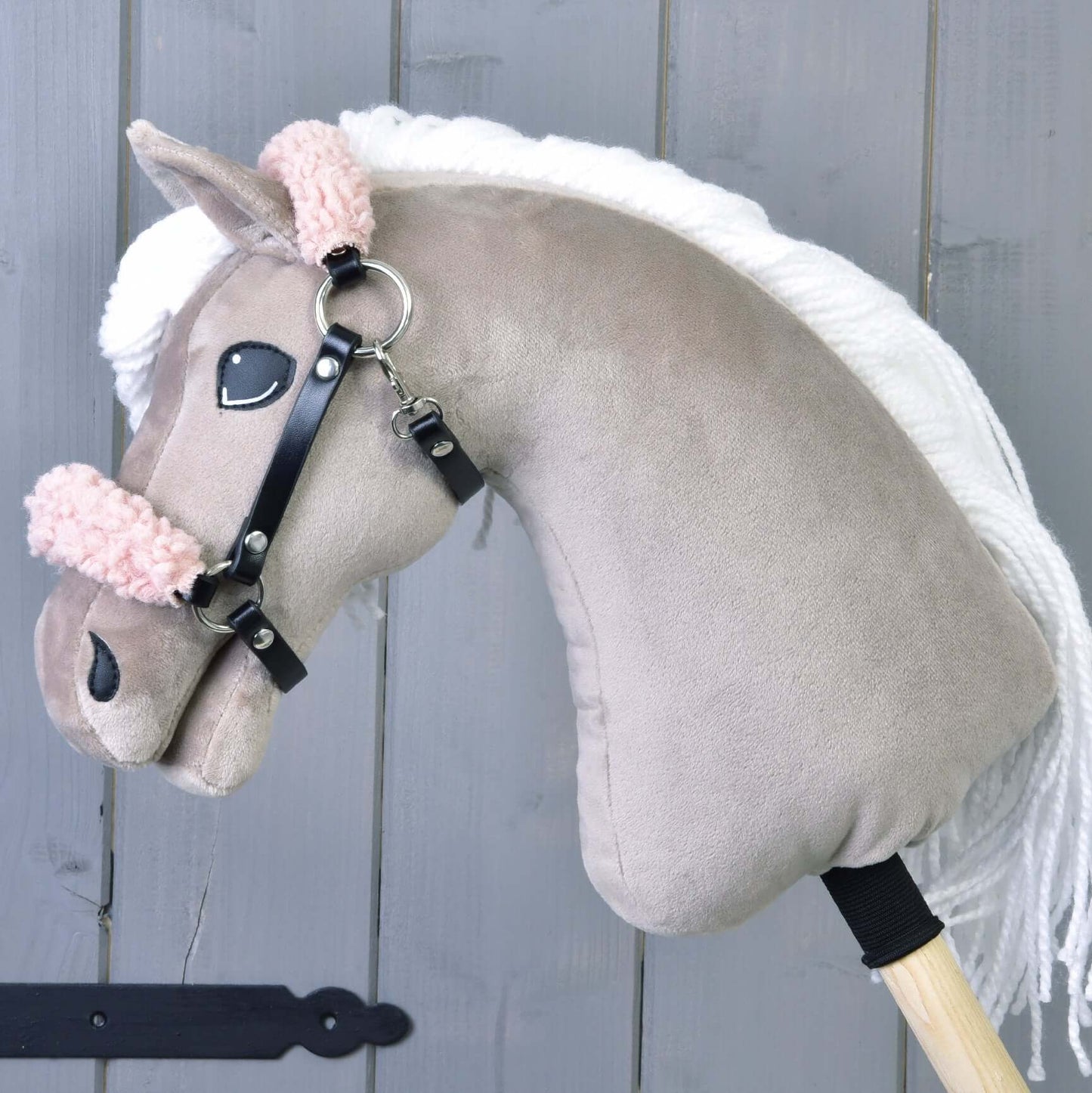 Licou noir avec fourrure vieux rose - similicuir de Hobby Horse LarDen sur un Hobby Horse en peluche à tête brun clair, crinière blanche, bridon rose duveteux et licou noir en similicuir, monté sur un bâton en bois devant un mur en bois gris.