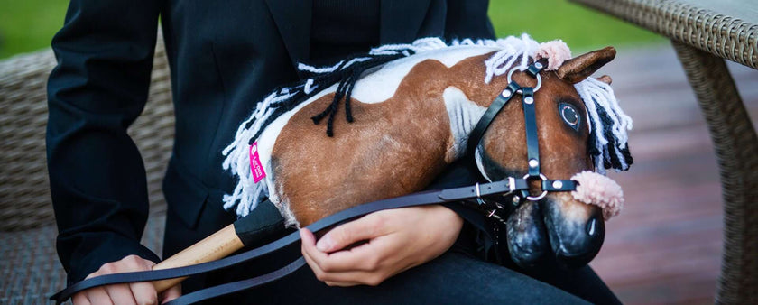 A person in a black blazer sits in a wicker chair, holding a brown and white Hobby Horse with a bridle and yarn mane on their lap, ready for some Hobby Horsing fun.
