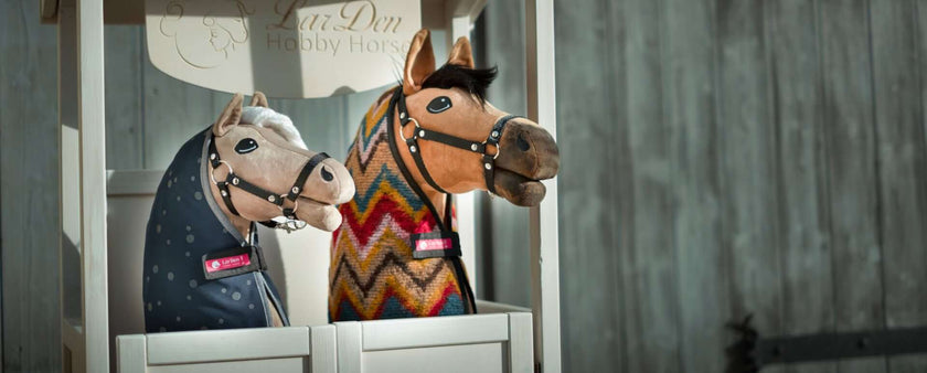 Two realistic hobby horse toys with bridles stand side by side in a wooden stall, perfect for hobby horsing fun. One features a light brown spotted design, while the other is tan with a colorful zigzag-patterned fabric neck. A wooden fence is in the background.