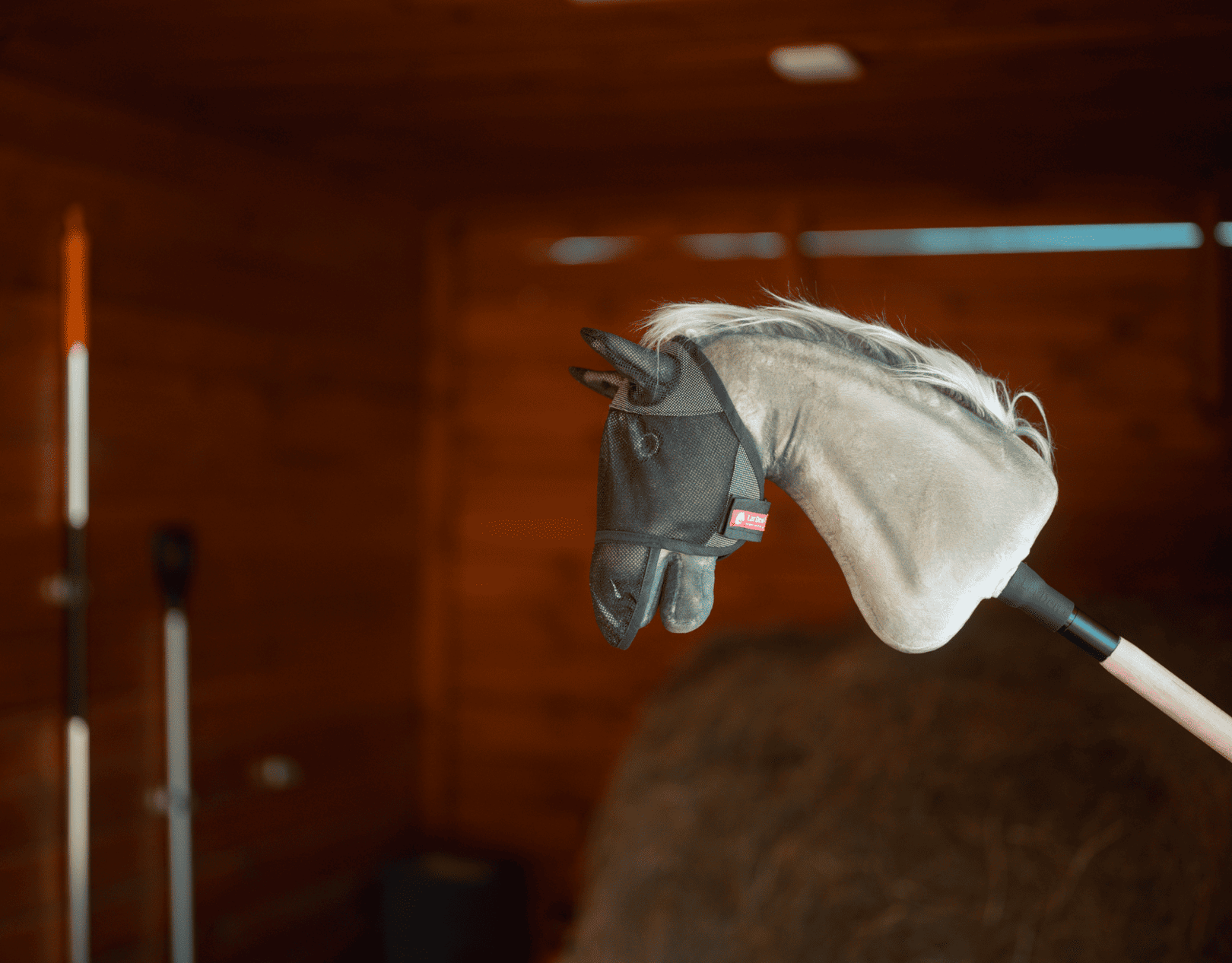 Ein Hobby Horse mit grauem Plüschkopf und schwarzem Zaumzeug ist auf einem Stock in einer Holzscheune montiert, umgeben von Hofwerkzeugen und Heu – ideal, um fantasievolle Hobby Horsing Abenteuer zu wecken.