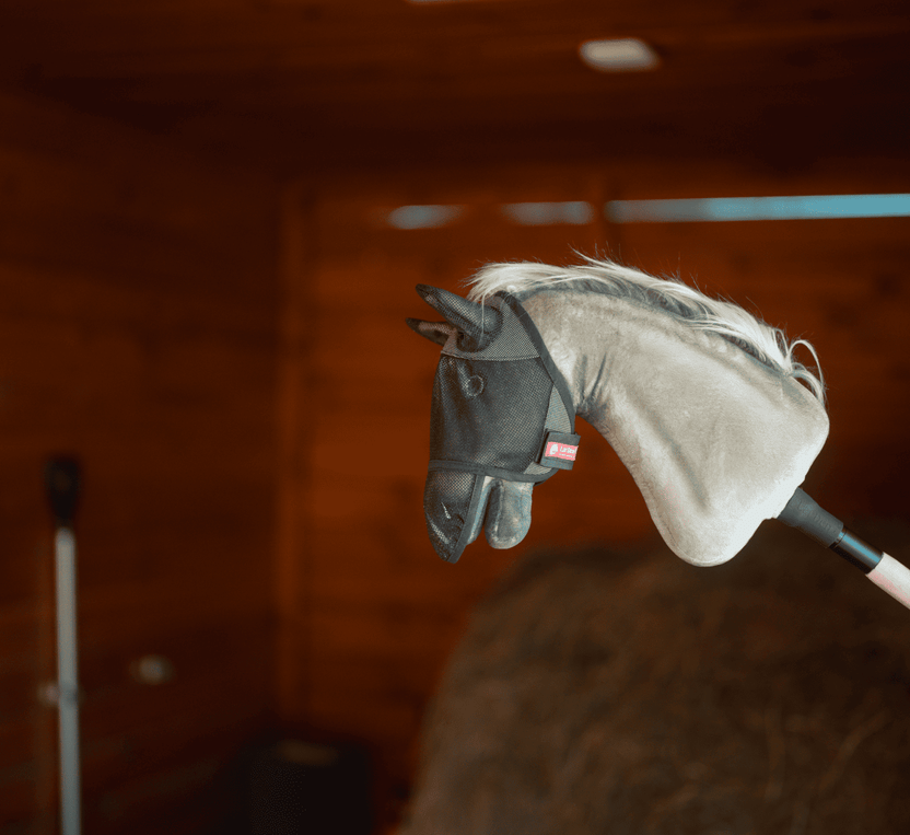 Ein Plüschkopf eines Hobby Horse auf einem Stock, mit schwarzer Fliegenmaske aus Mesh, ist in einem Innenraum mit Holzwänden und einem Heustapel zu sehen – ideal für fantasievollen Hobby Horsing Spaß.