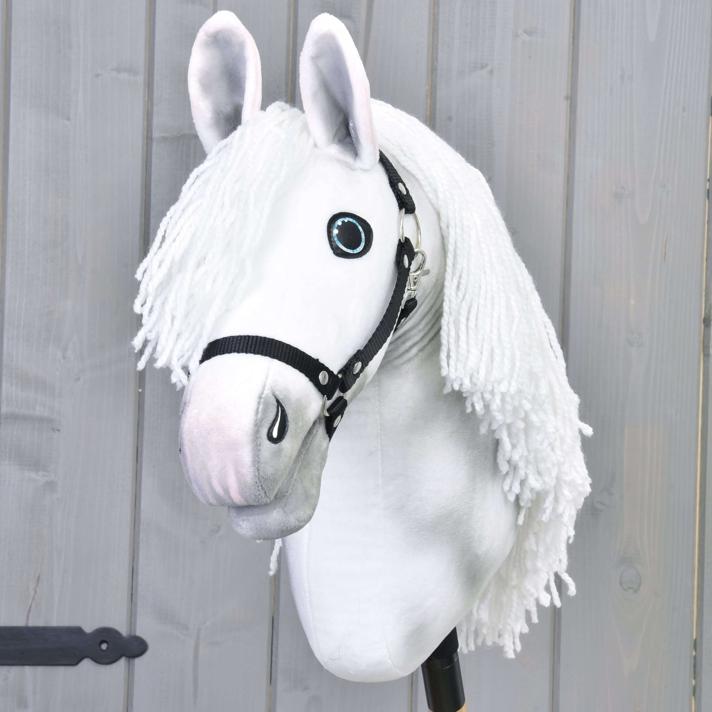 Hobby Horse Elektra avec licol noir de Hobby Horse LarDen, jument blanche peinte à la main avec longue crinière en fil, yeux bleus et licol noir, montée sur un bâton et présentée devant un mur en bois gris clair.