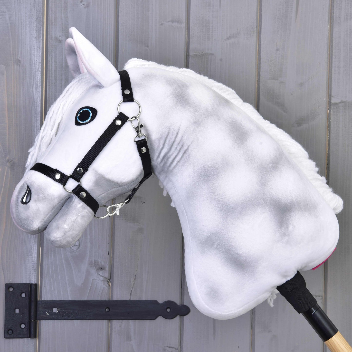 Hobby Horse Elektra avec licol noir de Hobby Horse LarDen, avec design blanc en peluche, yeux bleus et licol noir, monté sur un bâton, présenté ici devant un mur en bois gris.
