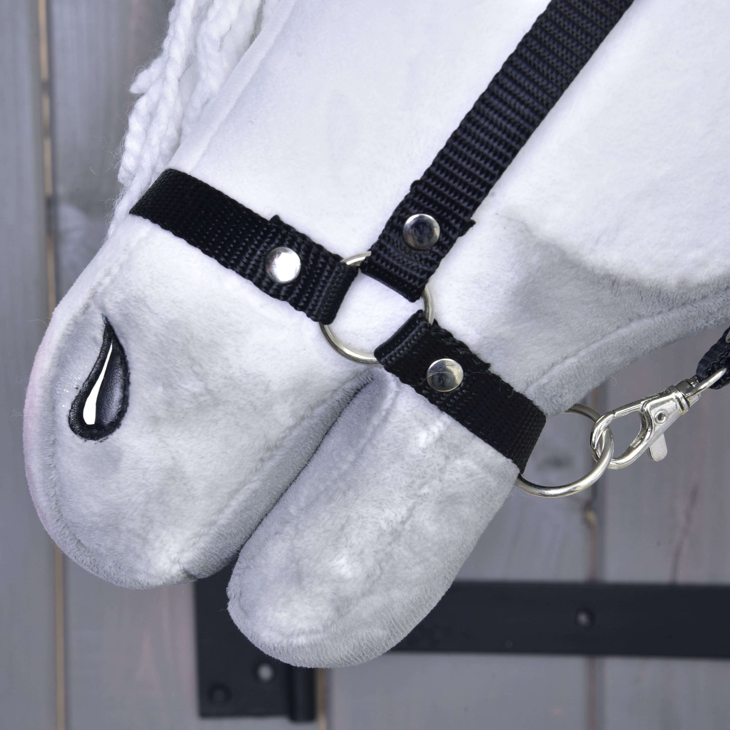 Gros plan sur Hobby Horse Elektra avec licol noir de Hobby Horse LarDen, avec visage blanc en peluche, licol en tissu noir avec anneaux et clip argentés, narine cousue en noir, sur fond en bois.