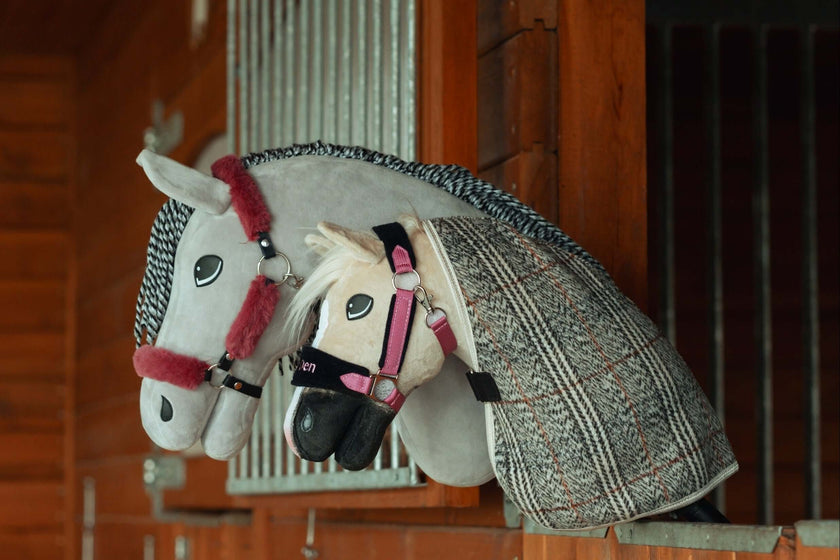 Zwei Hobby Horse Plüschspielzeuge mit Zaumzeug und Decken ruhen auf einer hölzernen Stalltrennwand und ähneln echten Pferden in einer Box. Ideal für Hobby Horsing Fans, die weißen und grauen Pferde haben detailreiche Merkmale und Ausrüstung.