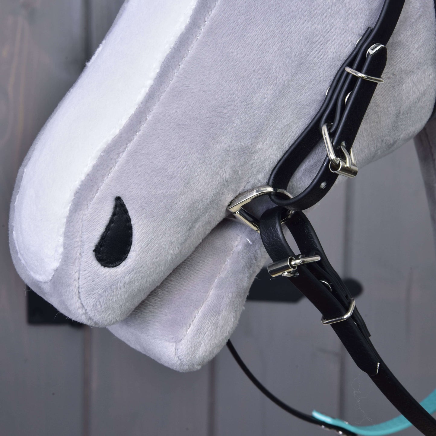Nahaufnahme eines grauen Hobby Horse von Hobby Horse LarDen mit Komplettes Zaumzeug ohne Nasenriemen Türkis und Zügeln, mit schwarzem, tropfenförmigem Auge vor unscharfem grauem Hintergrund.