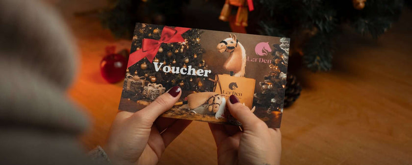 A person holding a festive voucher featuring cartoon dog illustrations, a red bow, and Christmas decorations next to a Christmas tree—perfect for any Hobby Horse enthusiast during the holiday season.