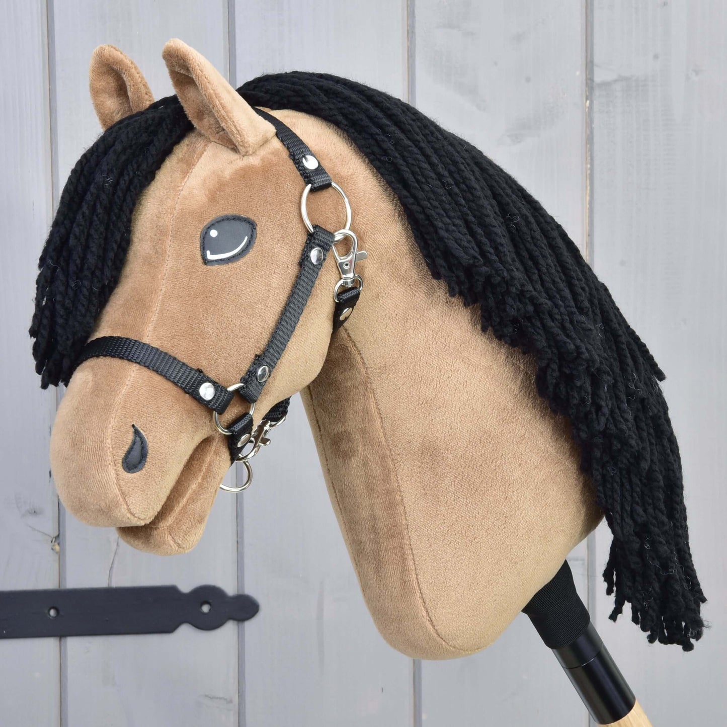 Hobby Horse Samantha mit Schwarzem Halfter von Hobby Horse LarDen zeigt einen hellbraunen Stoffkopf mit schwarzer Garnmähne und ist von einer englischen Vollblutstute inspiriert, auf einem Holzstab vor grauen Holzpaneelen.