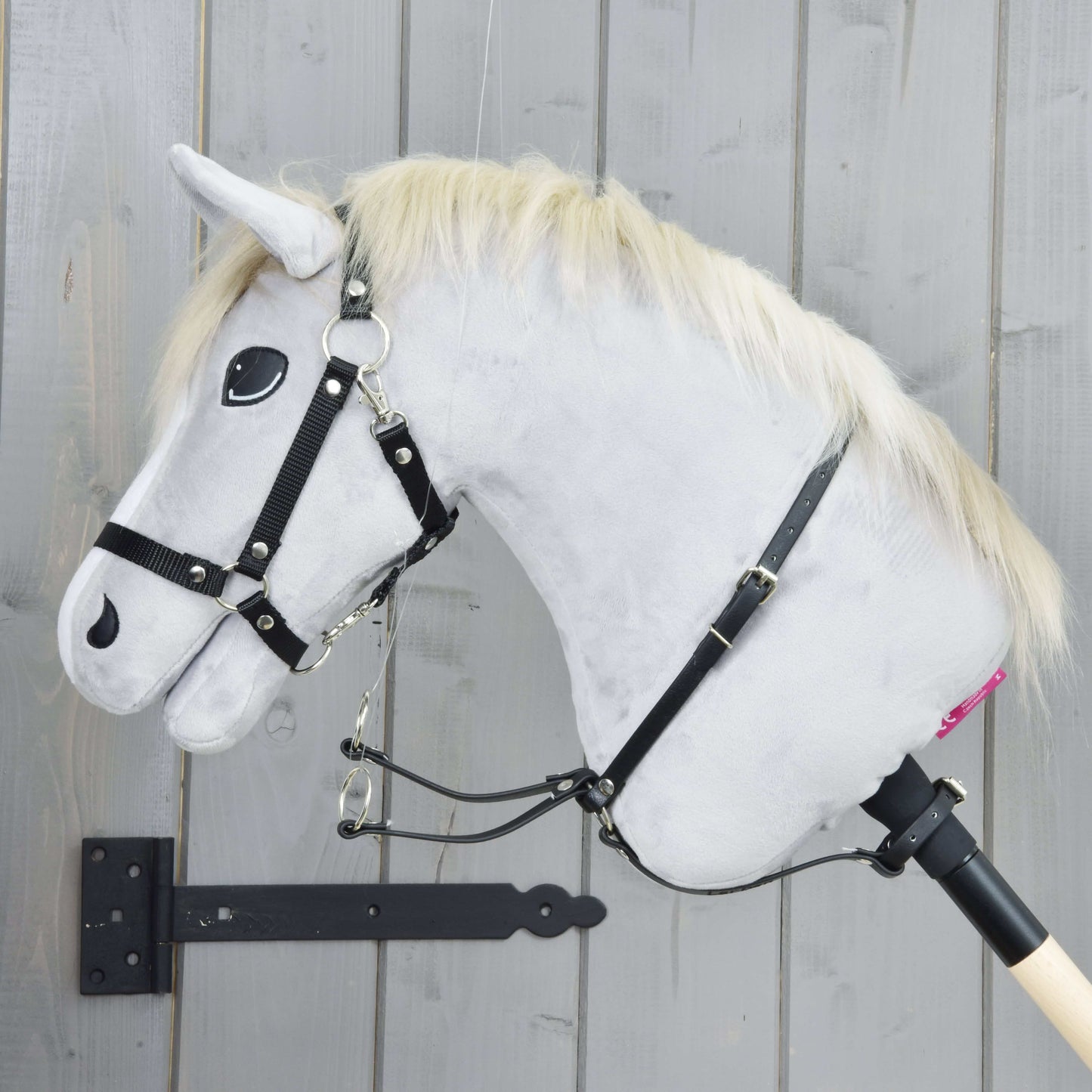 Martingale noir de Hobby Horse LarDen, Hobby Horse en peluche grise avec crinière blonde, bride noire, martingale et rênes, monté sur un bâton en bois devant un mur en bois gris clair.