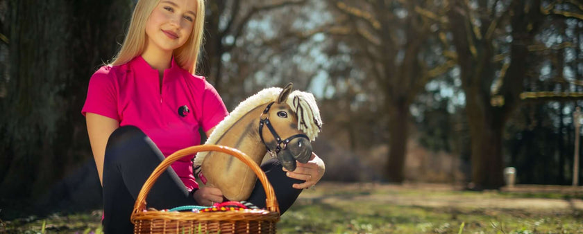 A young woman in a pink shirt sits outdoors on grass, smiling and holding a hobby horse beside a wicker basket filled with colorful items—capturing the playful spirit of hobby horsing amid sunlit trees and a blurred, peaceful background.