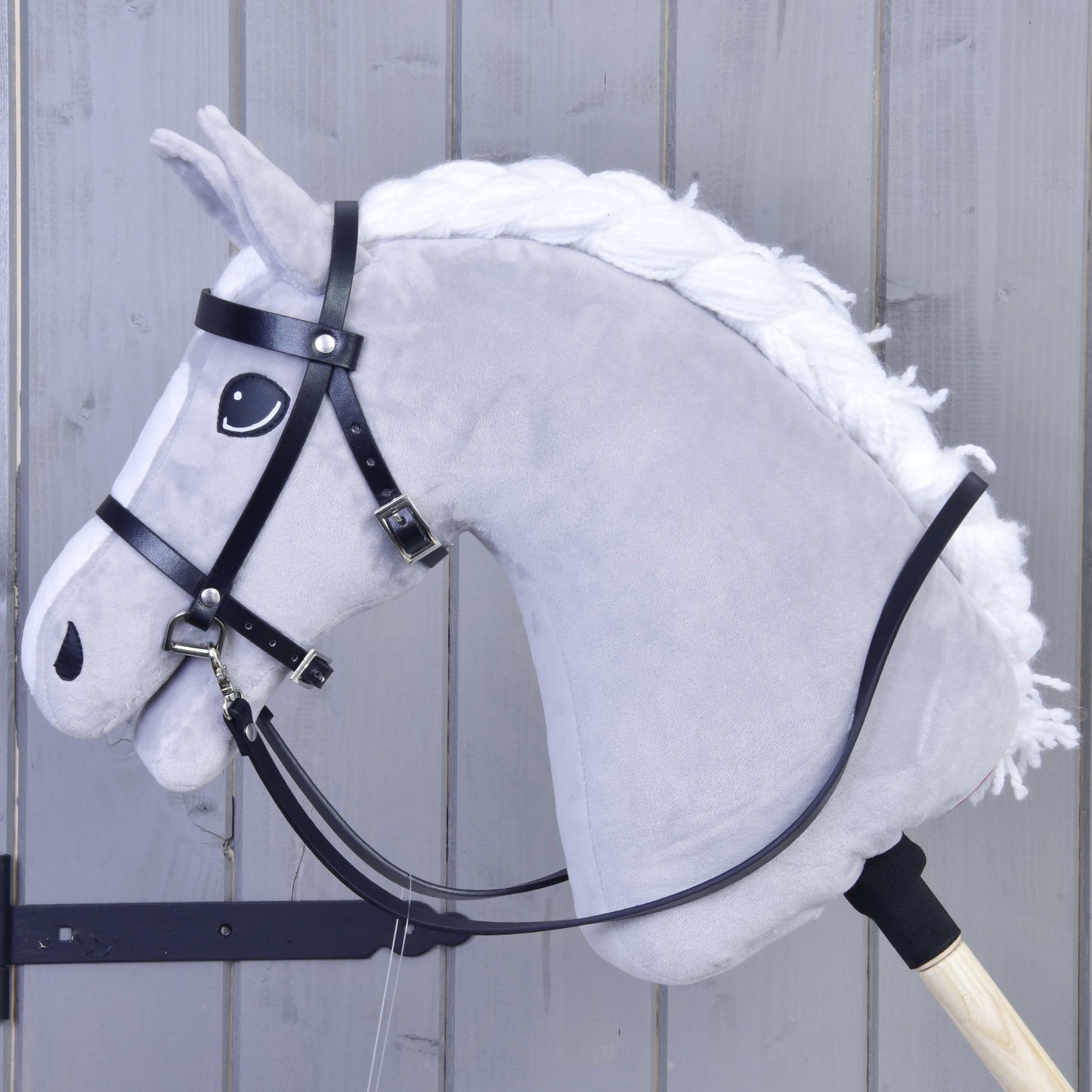 Nahaufnahme eines grauen Plüsch-Hobby Horse mit weißer Mähne auf Holzstab mit Schwarzes Zaumzeug aus Kunstleder von Hobby Horse LarDen, vor einem hellen Holzpaneel-Hintergrund.
