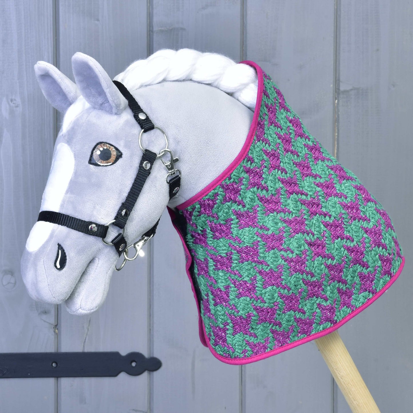Das Set Coco von Hobby Horse LarDen zeigt ein graues Hobby Horse mit schwarzem Zaumzeug, gestylt mit einem Deckenset mit Hahnentrittmuster in Lila und Grün, rosa Besatz und Fliegenhaube, vor einer grauen Holzwand.