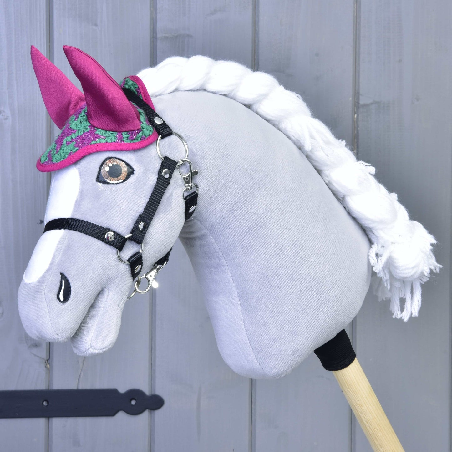 Das Hobby Horse LarDen Set Coco mit grauem Körper, weißer Mähne, schwarzem Zaumzeug und rosa Fliegenhaube steht vor einem grauen Holzhintergrund.