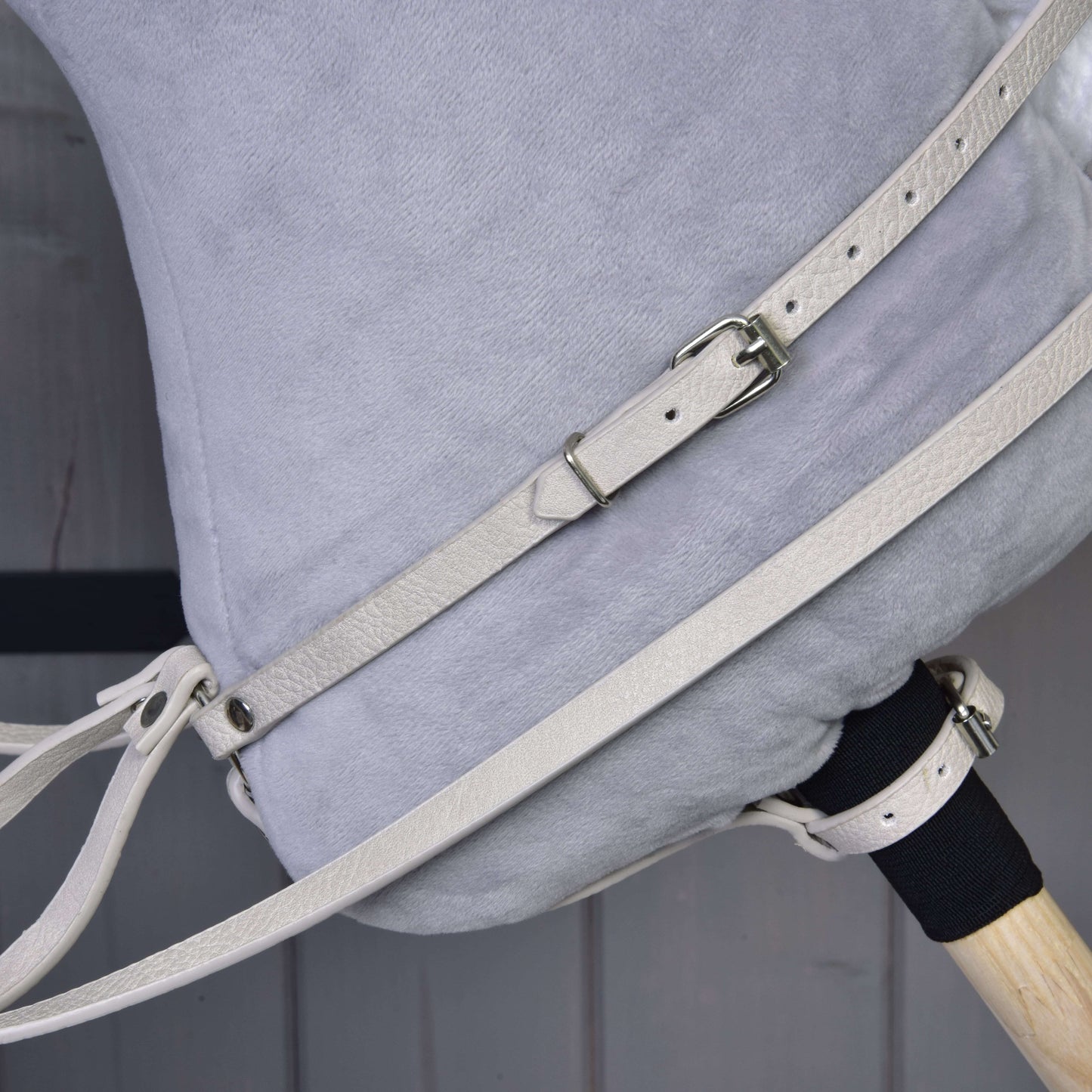 Gros plan sur le côté d’un Hobby Horse en peluche Hobby Horse LarDen, avec tissu gris clair et ensemble similicuir Aqua - bride, rênes et martingale avec boucles, ainsi qu’une partie de la poignée en bois, sur fond en bois.