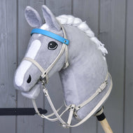 Hobby Horse LarDen mit grauem Plüsch-Hobby Horse mit geflochtener weißer Mähne auf einem Holzstab und dem Kunstlederset Aqua - Zaumzeug, Zügel und Martingale vor einer grauen Holzwand.